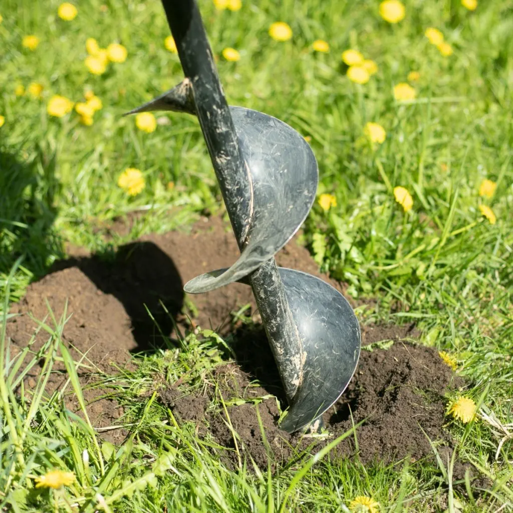 A drill auger bit that is breaking through new garden soil with grass and dandelions.