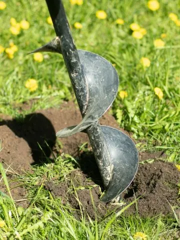 A drill auger bit that is breaking through new garden soil with grass and dandelions.